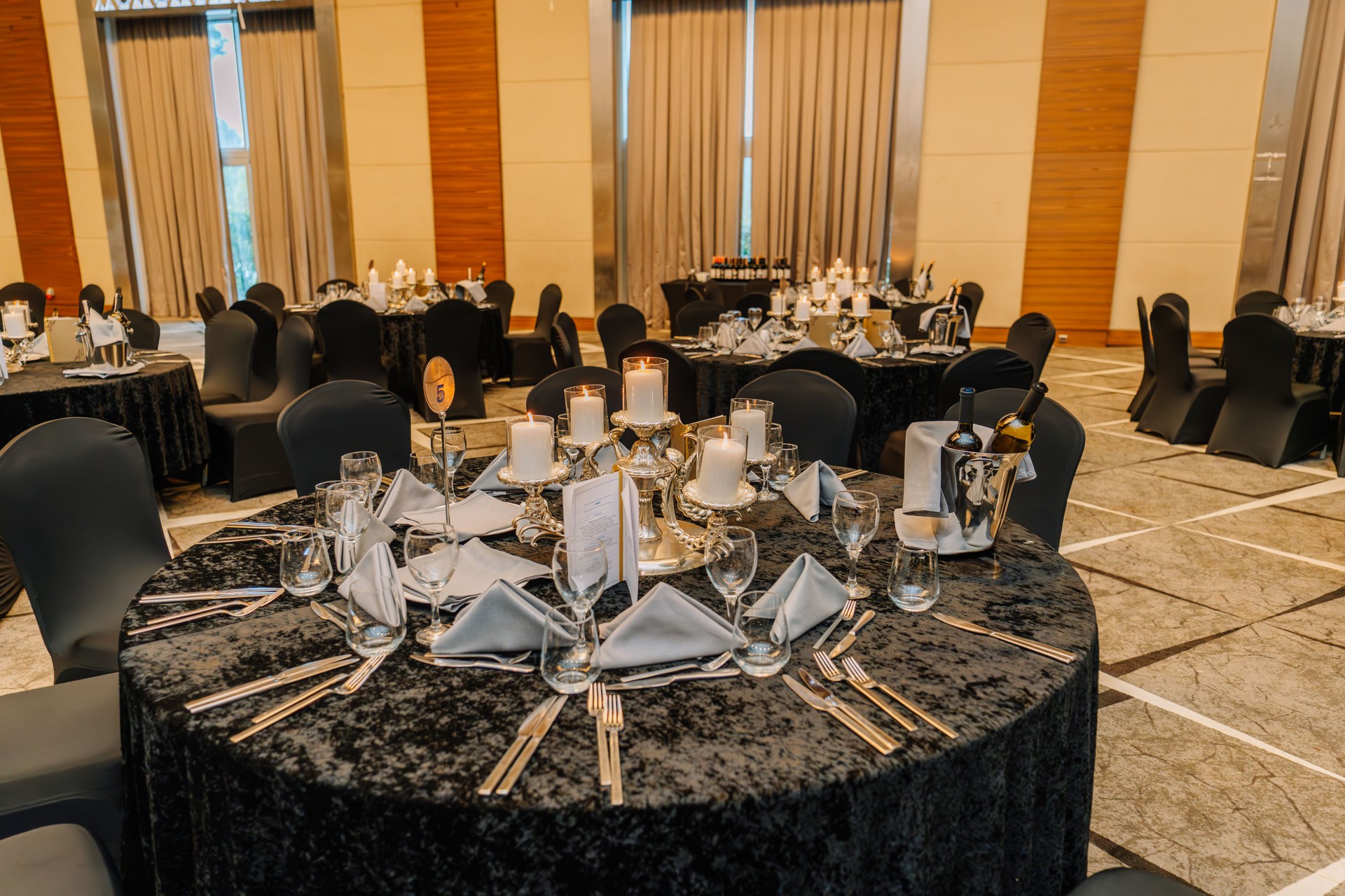 Luxurious banquet hall setup with round tables, candle centerpieces, wine bottles, and elegant place settings prepared for a formal dinner event.